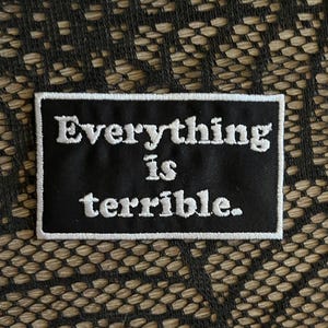 May include: A rectangular black embroidered patch with a white border. The text "Everything is terrible." is stitched in white thread in a serif font across the center of the patch, creating a high-contrast design with clear, legible lettering.