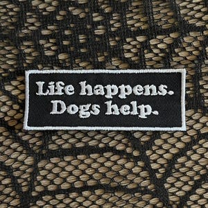 May include: A rectangular black embroidered patch with a white border. The text "Life happens. Dogs help." is stitched in white thread across the center of the patch in a clear, sans-serif font, creating a high-contrast design.