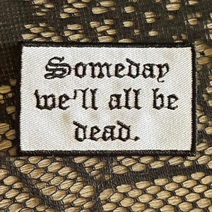 May include: A rectangular embroidered patch with a white background and a black border. The patch features black text in a gothic-style font that reads, Someday we'll all be dead. The embroidery is clean and the lettering is clearly legible.
