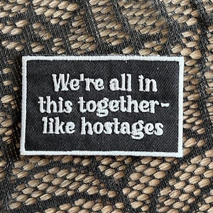 May include: A black rectangular patch with white embroidered text that reads "We're all in this together - like hostages." The patch has a white border and is set against a textured background.