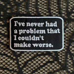 May include: A rectangular black embroidered patch with a white border and white stitched text that reads, I've never had a problem that I couldn't make worse. The patch features a clean, bold font and a durable fabric construction with finished edges.