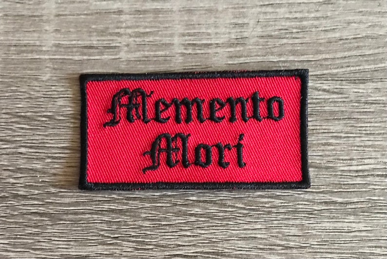 May include: Red rectangular embroidered patch with black stitching and the text "Memento Mori" in a gothic font.