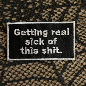May include: A rectangular black embroidered patch with a white border. The patch features the white embroidered text "Getting real sick of this shit." The lettering is in a clean, sans-serif font, centered on the black fabric background.