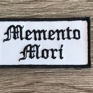 May include: White embroidered patch with black lettering that reads "Memento Mori" in an old English font.