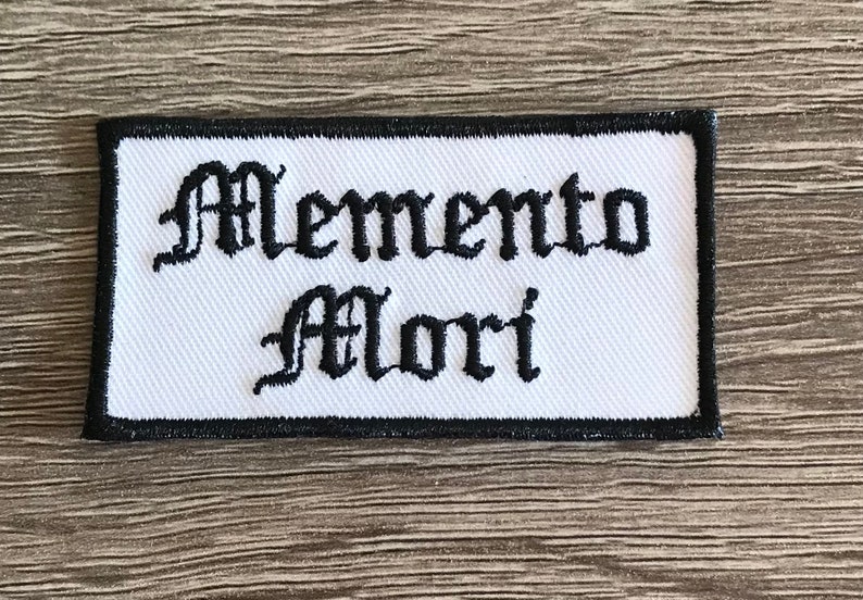 May include: White embroidered patch with black lettering that reads "Memento Mori" in an old English font.