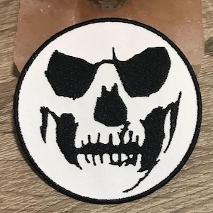 May include: White circular patch with a black embroidered skull design. The skull has a simple design with two eye sockets, a nose, and a mouth with teeth.