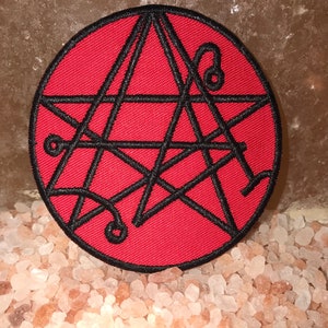 May include: A red embroidered patch with a black pentagram design. The pentagram is surrounded by a circle and has a complex pattern of lines and curves.