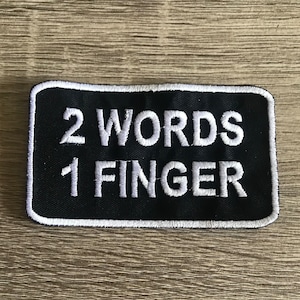 May include: Black embroidered patch with white stitching. The patch says "2 WORDS 1 FINGER".