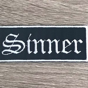 May include: Black and white embroidered patch with the word "Sinner" in a gothic font.