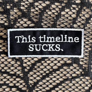 May include: A rectangular black embroidered patch with a white border and the white text "This timeline SUCKS." The patch is positioned against a black lace fabric background with a repeating hexagonal and floral pattern throughout the material.