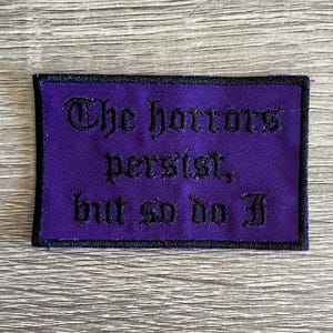 Puede incluir: Parche bordado morado con texto negro que dice "The horrors persist, but so do I."