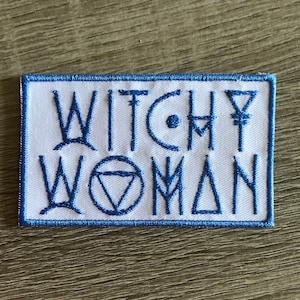 May include: A rectangular embroidered patch with a white background and a blue border. The text WITCHY WOMAN is stitched in blue thread, featuring stylized lettering where the letter O is replaced by a circle containing an inverted triangle.