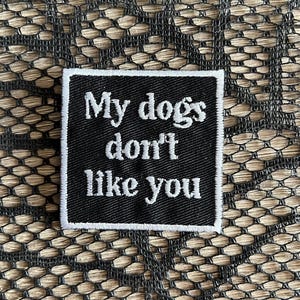 May include: A square, black and white embroidered patch with the text "My dogs don't like you". The patch has a white border and is set against a textured background.