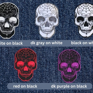 May include: Five embroidered iron-on patches featuring a sugar skull design in various color combinations. The skull designs are black with white, white with dark gray, black with white, black with red, and black with dark purple.