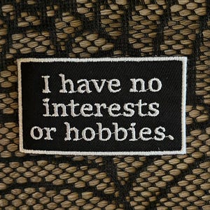 May include: A rectangular black embroidered patch with a white border. The patch features the white stitched text "I have no interests or hobbies." in a clean, sans-serif font, centered against the black fabric background of the patch.