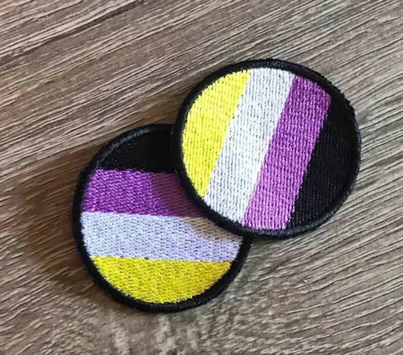 May include: Two round embroidered patches with the colors of the non-binary pride flag. The top patch has yellow, white, purple, and black stripes. The bottom patch has yellow, white, purple, and black stripes in a different order.