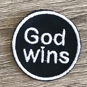 Puede incluir: Un parche circular negro y blanco con costuras blancas alrededor del borde. El parche tiene las palabras "God Wins" escritas en blanco sobre un fondo negro.