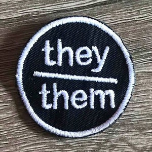 May include: Black and white embroidered patch with the text "they/them" in white lettering.