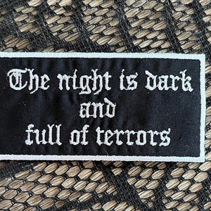 May include: Black and white embroidered patch with the text "The night is dark and full of terrors".