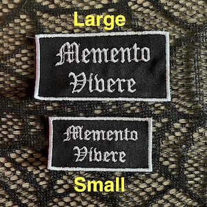 May include: Two black rectangular patches with white embroidered text. The larger patch reads "Large" above the words "Memento Vivere". The smaller patch reads "Small" below the same text.
