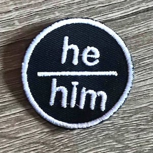 May include: Black and white embroidered patch with the words "he/him" in a circle.