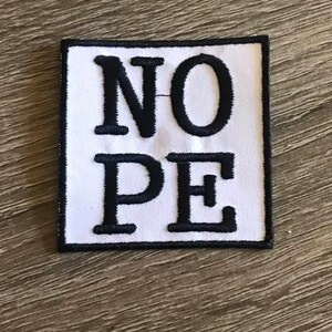May include: White patch with black embroidered text that reads "NOPE".