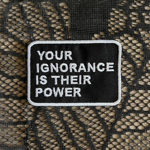 May include: A rectangular black embroidered patch with a white border. The patch features the white text "YOUR IGNORANCE IS THEIR POWER" in a bold, sans-serif font. The design is centered and clearly legible against the black fabric background.