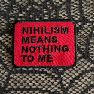 May include: A rectangular embroidered patch with a vibrant red background and a black border. The text "NIHILISM MEANS NOTHING TO ME" is stitched in bold, black, sans-serif capital letters across the center of the patch surface.