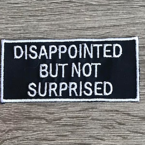 May include: Black embroidered patch with white text that reads "DISAPPOINTED BUT NOT SURPRISED".
