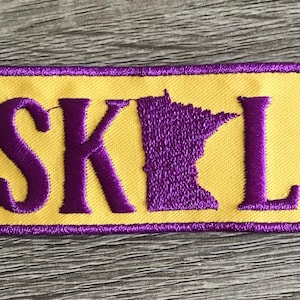 May include: Purple and yellow embroidered patch with the word "SKOL" and an outline of the state of Minnesota.