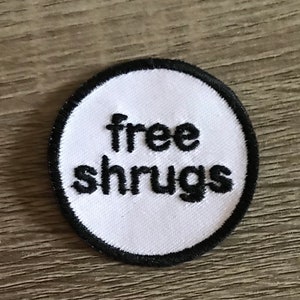 May include: White round embroidered patch with black stitching. The patch says "free shrugs" in black.