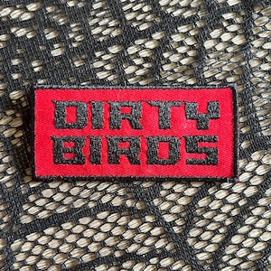 May include: A rectangular patch with the words "DIRTY BIRDS" embroidered in black on a red background. The patch is likely for clothing or accessories.