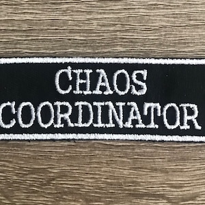 May include: A black rectangular patch with white embroidered text that reads "CHAOS COORDINATOR". The patch has a white border and is set against a wooden background. This patch is suitable for jackets, bags, or other fabric items.