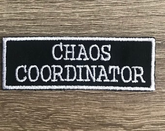 May include: A black rectangular patch with white embroidered text that reads "CHAOS COORDINATOR". The patch has a white border and is set against a wooden background. This patch is suitable for jackets, bags, or other fabric items.