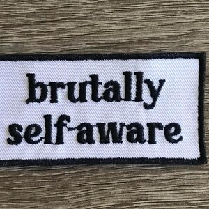 Puede incluir: Parche blanco bordado con costuras negras. El parche dice "brutally self-aware".