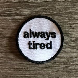 May include: White round patch with black embroidered text that reads "always tired".