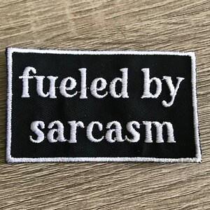 May include: A black rectangular patch with white embroidered text that reads "fueled by sarcasm." The patch has a white border and is displayed on a light wood surface.