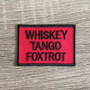 May include: A rectangular patch featuring the embroidered text "WHISKEY TANGO FOXTROT" in black on a red field. The patch is edged with a black border, suitable for attachment to fabric items.
