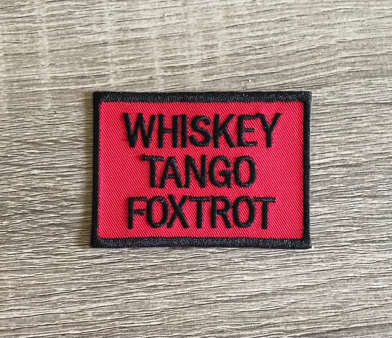 May include: A rectangular patch featuring the embroidered text "WHISKEY TANGO FOXTROT" in black on a red field. The patch is edged with a black border, suitable for attachment to fabric items.