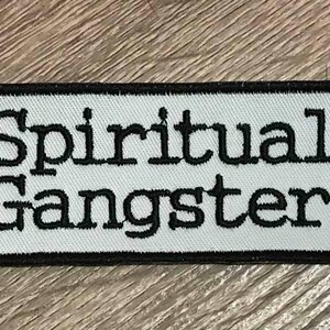 May include: A rectangular patch with the words "Spiritual Gangster" embroidered in black on a white background. The patch has a black border and is set against a wood grain surface.