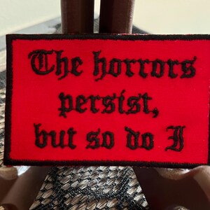Puede incluir: Parche de tela roja con texto bordado en negro que dice "The horrors persist, but so do I."