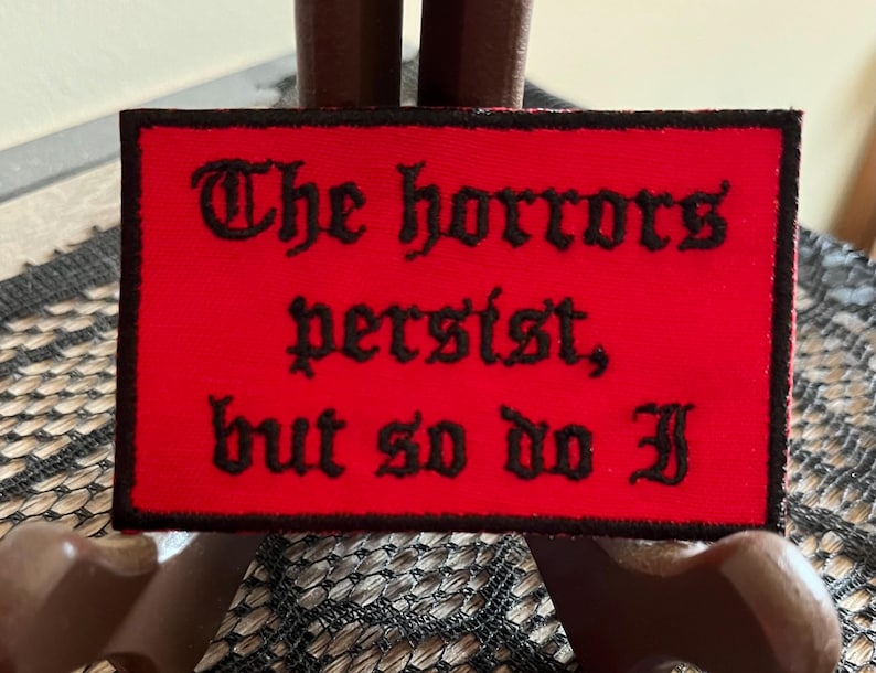 Puede incluir: Parche de tela roja con texto bordado en negro que dice "The horrors persist, but so do I."