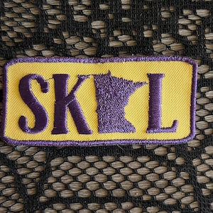 May include: A rectangular embroidered patch with a yellow background and a purple border. The patch features the purple letters S, K, and L, with the letter I replaced by a purple silhouette of the state of Minnesota, spelling out SKIL.