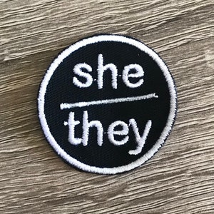 May include: A black and white embroidered patch with the words "she/they" in white.