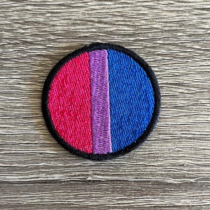 May include: A round embroidered patch with a pink, purple, and blue striped design. The patch is surrounded by a black border.