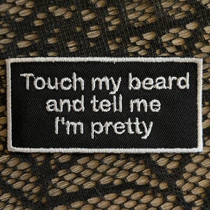 Puede incluir: Un parche rectangular negro bordado con un borde blanco y texto blanco cosido que dice: Touch my beard and tell me I'm pretty. El parche presenta una textura de tela densa y está diseñado para fijarse en prendas de vestir o accesorios.