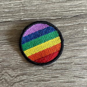 May include: A rainbow-colored embroidered patch in the shape of a circle. The patch has a black border and is made of fabric.