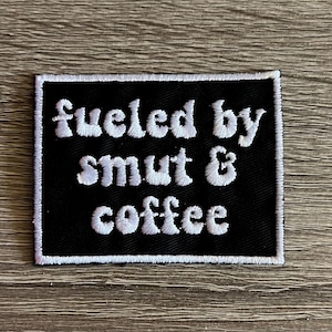 May include: A black rectangular patch with white embroidered text that reads "fueled by smut & coffee". The patch has a white border and is displayed on a wooden surface.