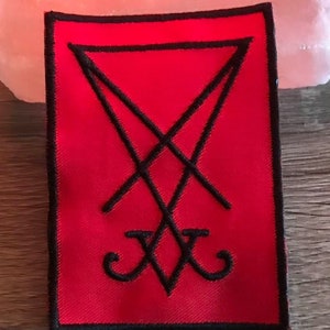 Puede incluir: Un parche rojo bordado con un símbolo de pentagrama invertido negro. El parche tiene un borde negro.