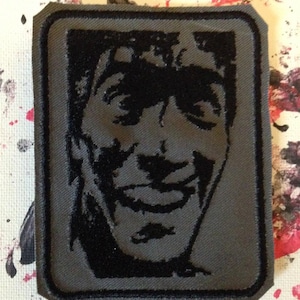 May include: A black and gray embroidered patch featuring a smiling face with a black background. The patch is sewn onto a gray fabric.
